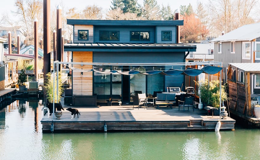 A two story modern turquoise home with a tin roof, outdoor deck with an awning and a dog at Island Cove Floating homes Marin in Portland Oregon